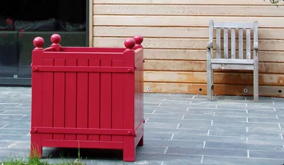Roter Orangerie Pflanzkübel aus lackiertem Akazienholz steht auf einer Terrasse neben einem Holzstuhl im Freien