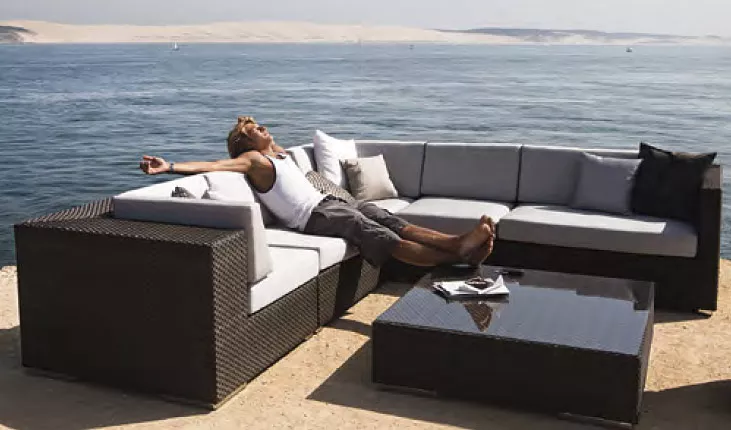 Ein Mann entspannt sich auf einer Dedon Lounge Gartenmöbelgarnitur am Meer mit einem Couchtisch und einem Buch auf einem sonnigen Tag.