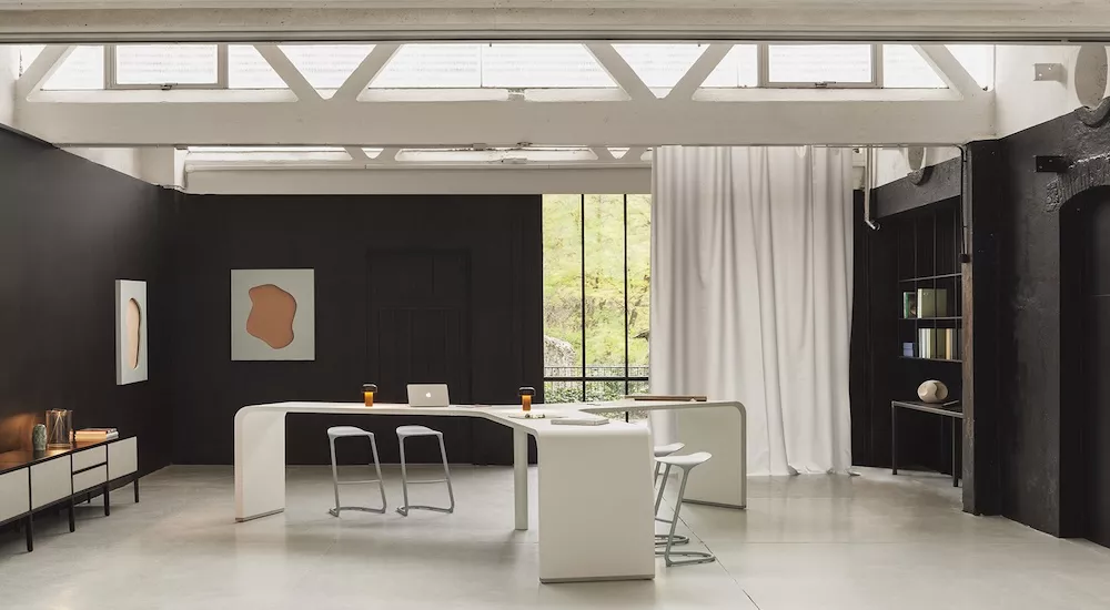 Heller Raum mit schwarzer Wand, weißem Tisch und Stühlen, Kunstwerken an der Wand und großen Fenstern, die Tageslicht hereinlassen.