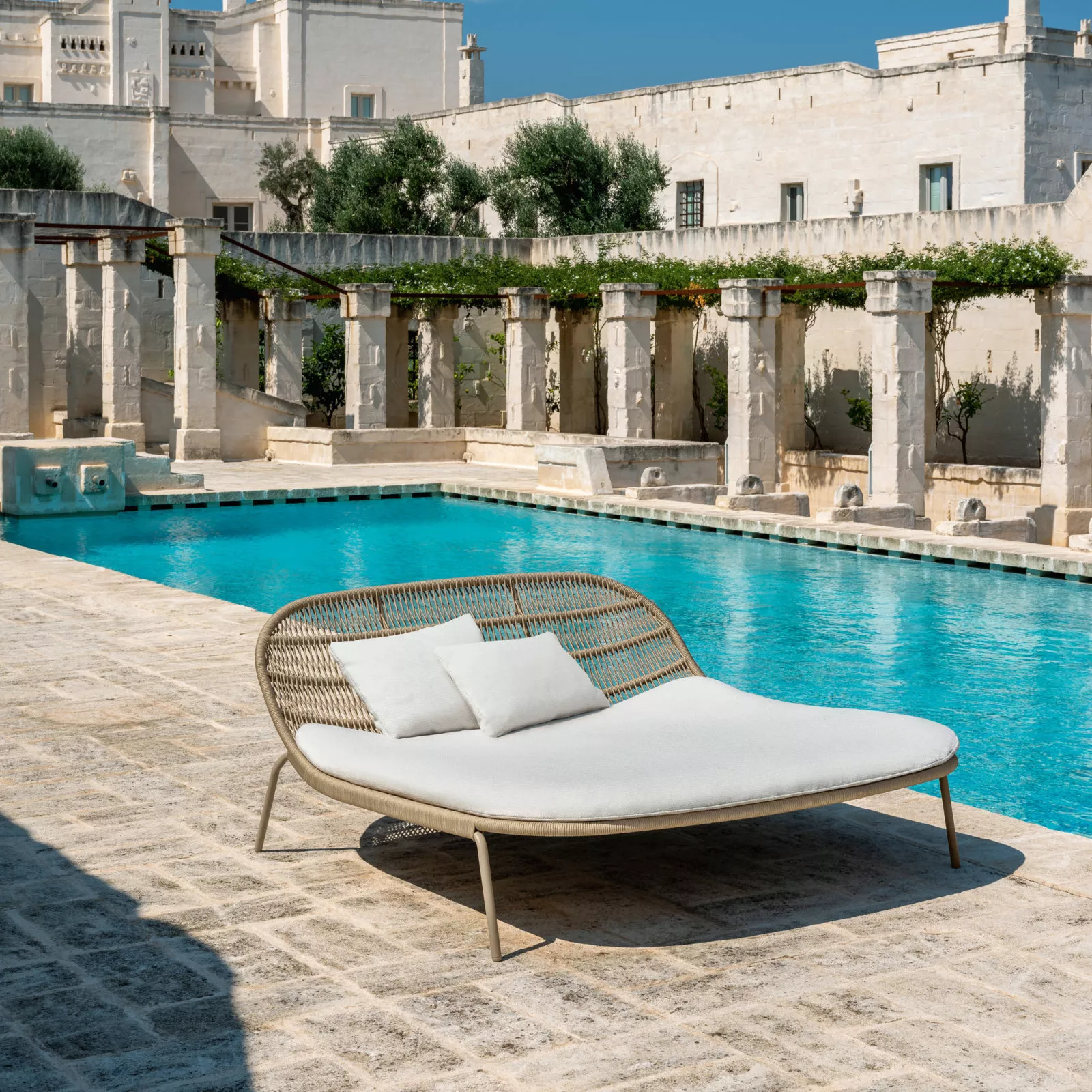 Alenti Panama Daybed in Beige mit Auflage, aufgestellt auf einer sonnigen Terrasse neben einem Pool, umgeben von antiker Architektur.