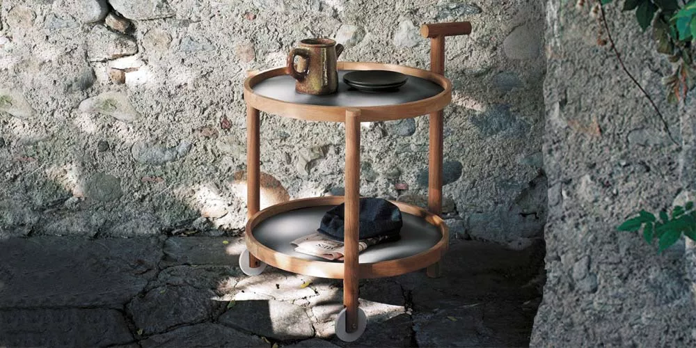 Roda Caddy Servierwagen aus Holz und Metall in Naturfarbe auf Steinboden vor einer rustikalen Steinwand, mit Tasse und Teller dekoriert.