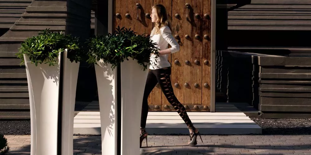 Frau geht an zwei weißen Vondom Alma Pflanzenkübeln vorbei, die vor einer Holztür auf einem gepflasterten Weg stehen.