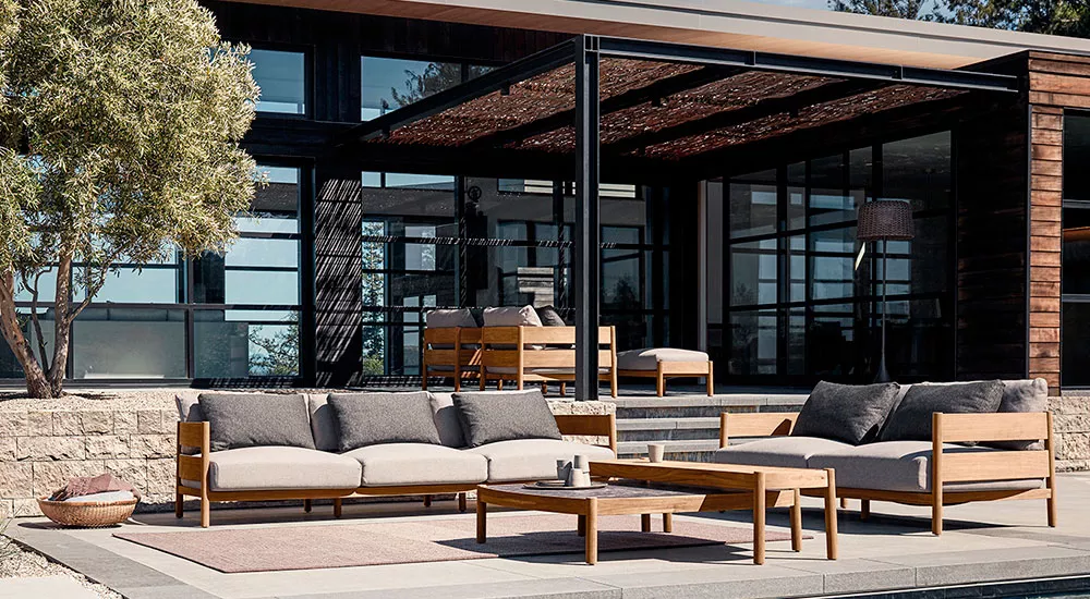 Gloster Haven Loungegruppe in Naturholz und Grau auf moderner Terrasse vor einem Haus mit großen Glasfenstern, entspannte Atmosphäre.