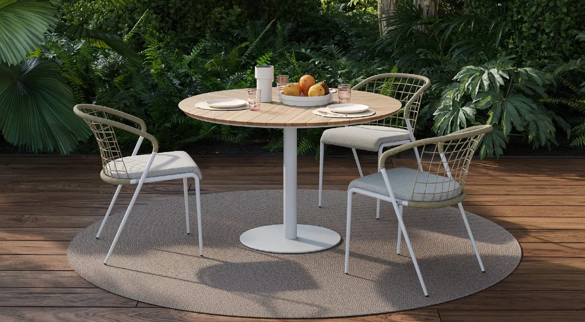 Runder Holztisch mit weißen Metallstühlen auf einem Teppich, umgeben von üppigem Grün im Garten, mit gedecktem Tisch für zwei Personen.