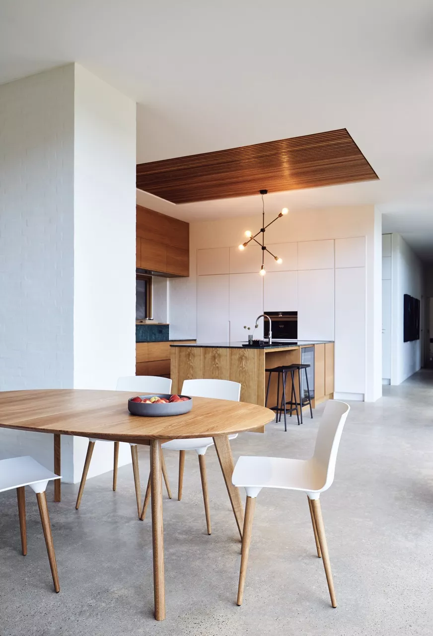 Heller Raum mit einem runden DK10 Holztisch und weißen TAC Stühlen von Villa Schmidt vor einer modernen Küche.