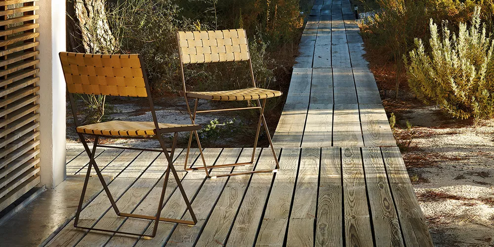 Zwei Andreu World Brisa Stühle in Holzoptik auf einer Holzterrasse in einem natürlichen Garten bei Sonnenuntergang.