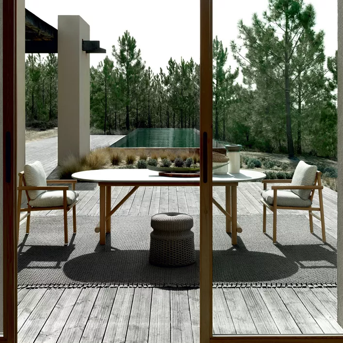 Zwei B&B Italia Ayana Stühle mit Armlehnen stehen an einem runden Tisch auf einer Terrasse mit Blick auf einen Pool und Bäume im Hintergrund