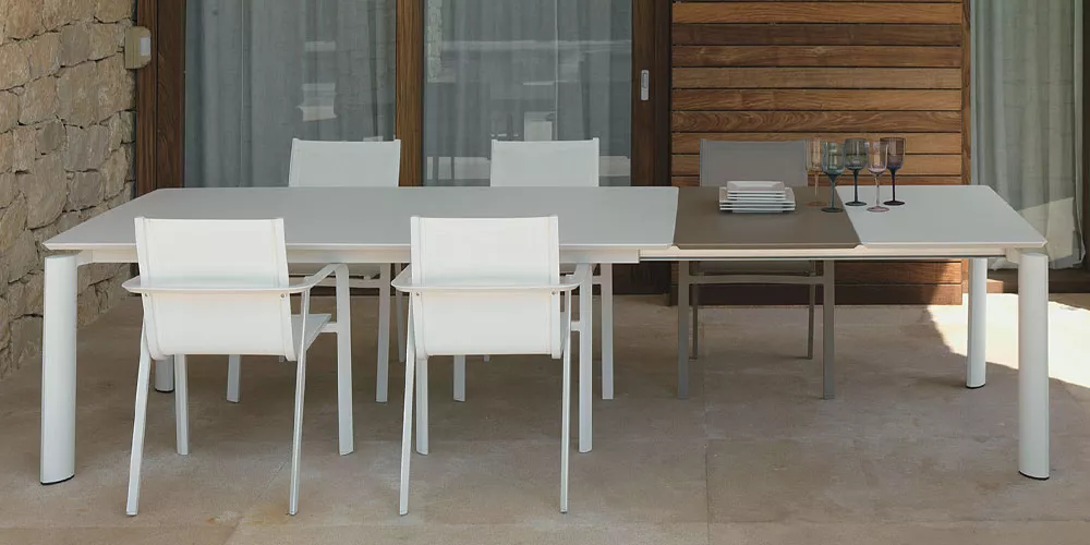 Talenti Milo Kollektion: Moderner Esstisch mit weißen Stühlen auf einer Terrasse vor einer Stein- und Holzwand.