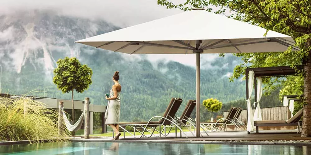 Glatz Palazzo Noblesse Sonnenschirm in Weiß, aufgestellt in einer idyllischen Berglandschaft neben einem Pool, mit Liegestühlen im Hintergrund.