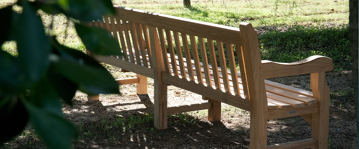 Teakholzbank von Villa Schmidt in natürlicher Umgebung, seitliche Ansicht, sonnendurchfluteter Hintergrund mit Bäumen und Gras.