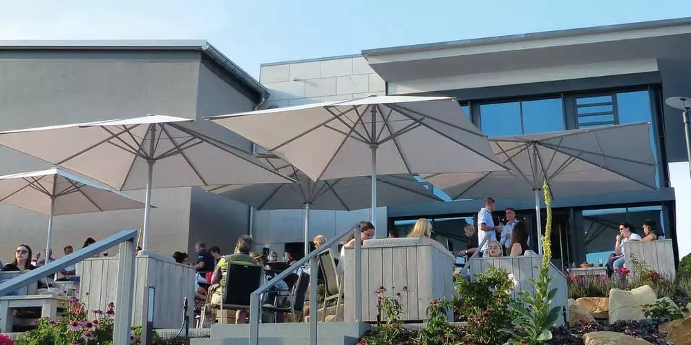 May Sonnenschirme Filius FS in Beige auf einer Terrasse mit Menschen, die unter den Schirmen entspannen, vor einem modernen Gebäude.
