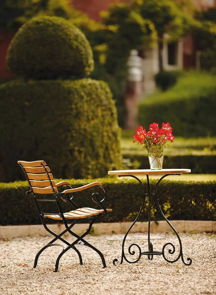 Schmiedeeiserner Tisch Classic in Schwarz mit Holzstuhl und Blumenstrauß auf Kiesweg vor gepflegtem Garten im Hintergrund.