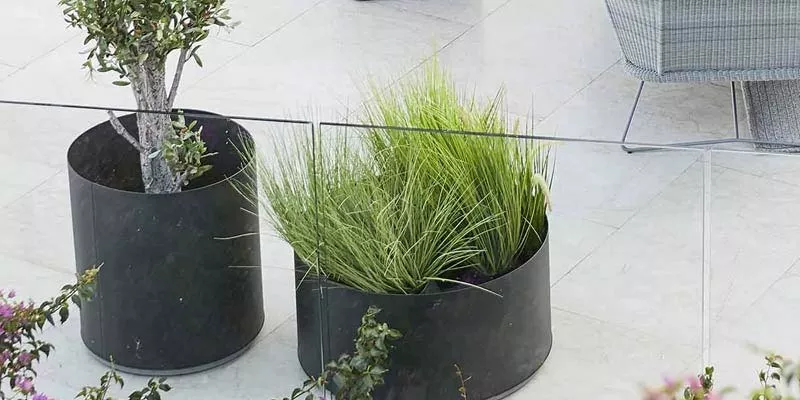 Zwei schwarze Cane-line Grow Pflanzgefäße auf einer Terrasse, eines mit einem Baum und das andere mit Gras, auf hellem Steinboden.