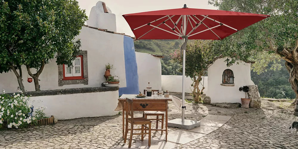 Glatz Fortero Sonnenschirm in Rot auf einer mediterranen Terrasse mit rustikalem Tisch und Stühlen, umgeben von weiß getünchten Gebäuden.