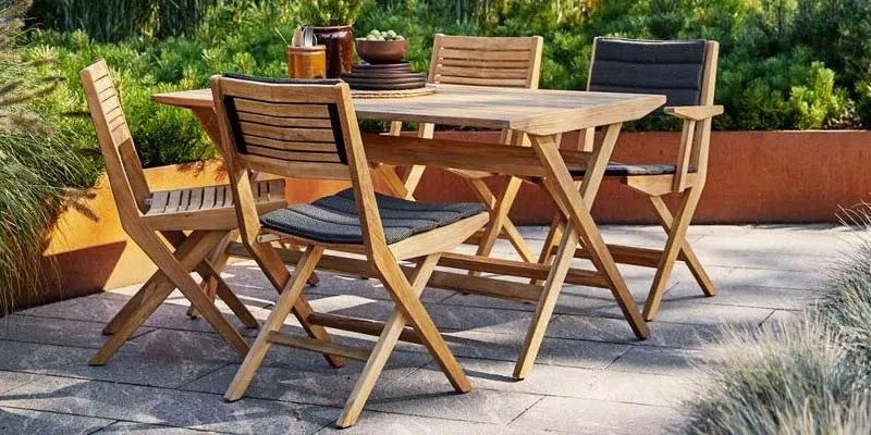 Cane-line Flip Essgruppe aus hellem Holz mit vier Stühlen und einem Tisch auf einer Terrasse, umgeben von grünen Pflanzen.