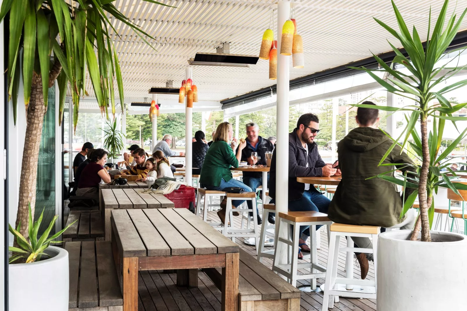 Außenbereich eines Cafés mit Bromic Smart-Heat Platinum SII Electric 2300W in Schwarz an der Decke, umgeben von Pflanzen und Gästen.