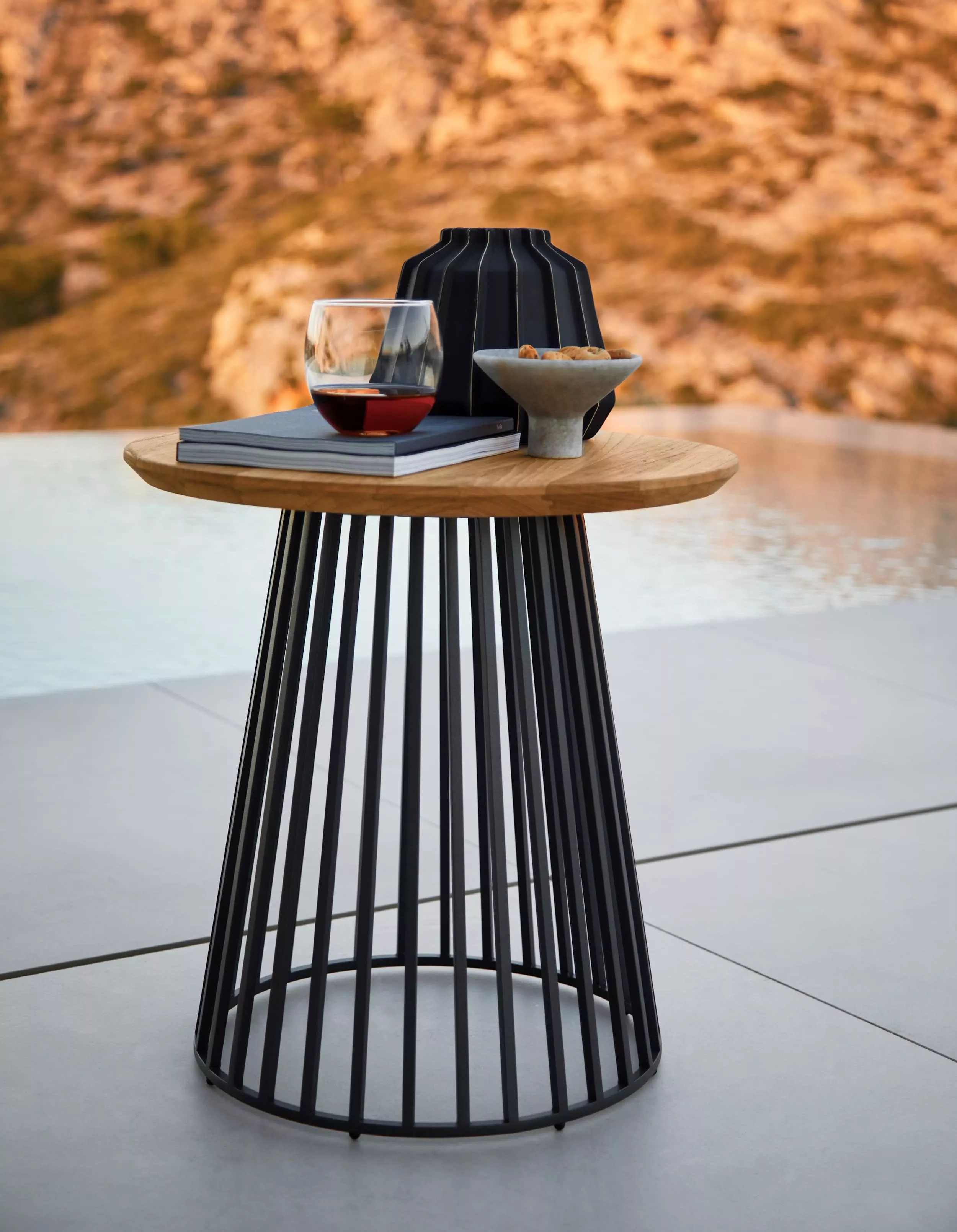 Solpuri Grid Beistelltisch mit einem Buch einem Glas Rotwein und einer Schale mit Snacks auf einer Terrasse mit Blick auf eine felsige Landschaft