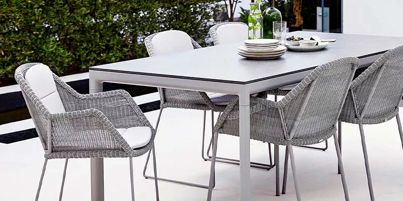 Cane-line Breeze Essgruppe in Grau auf einer Terrasse mit grünem Hintergrund, bestehend aus einem Tisch und mehreren Stühlen.