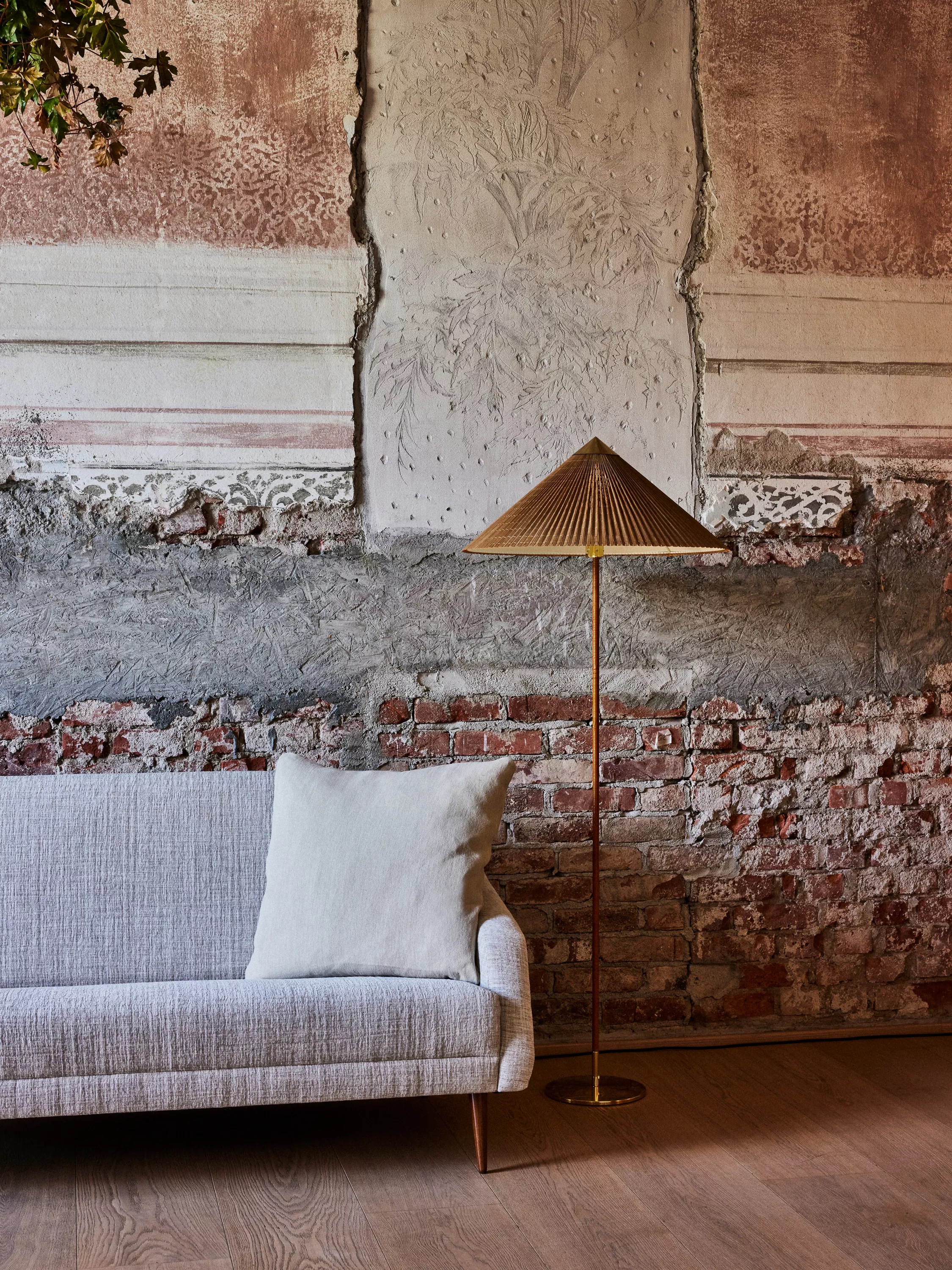 GUBI Stehleuchte mit Schirm steht neben einem grauen Sofa mit Kissen vor einer rustikalen Wand in einem gemütlichen Wintergarten.