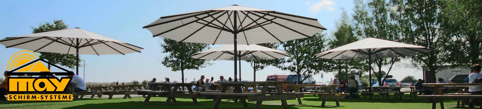 MAY Schirm-Systeme Sonnenschirme in Weiß auf einer Wiese mit Picknicktischen und Bäumen im Hintergrund bei sonnigem Wetter.
