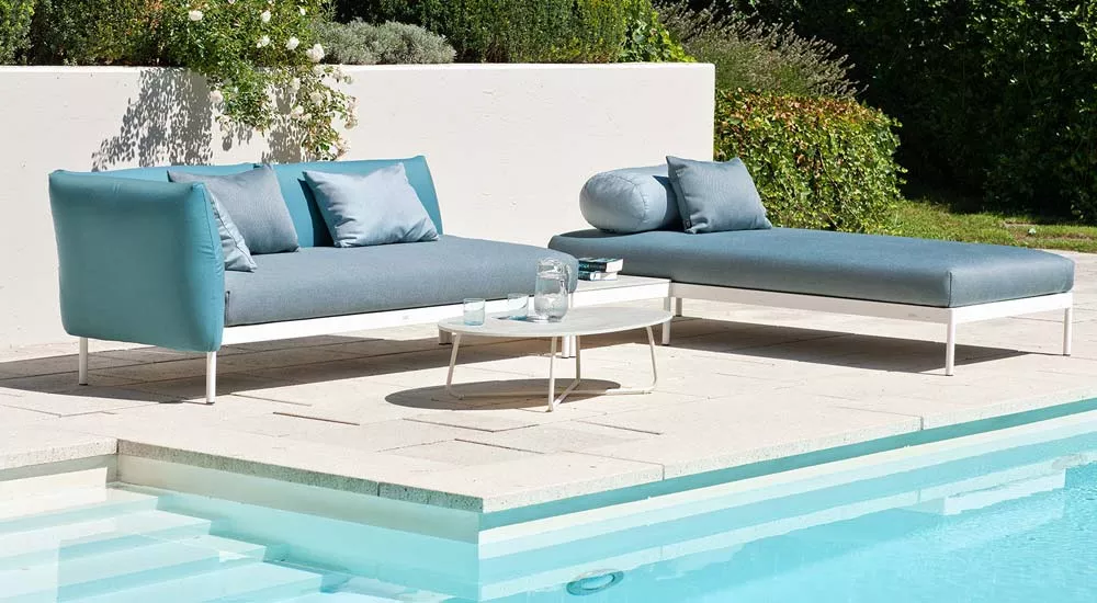 Fischer Möbel Kairos Loungegruppe in Blau auf heller Terrasse am Pool, bestehend aus Sofa und Liege mit weißem Gestell, umgeben von Grün.