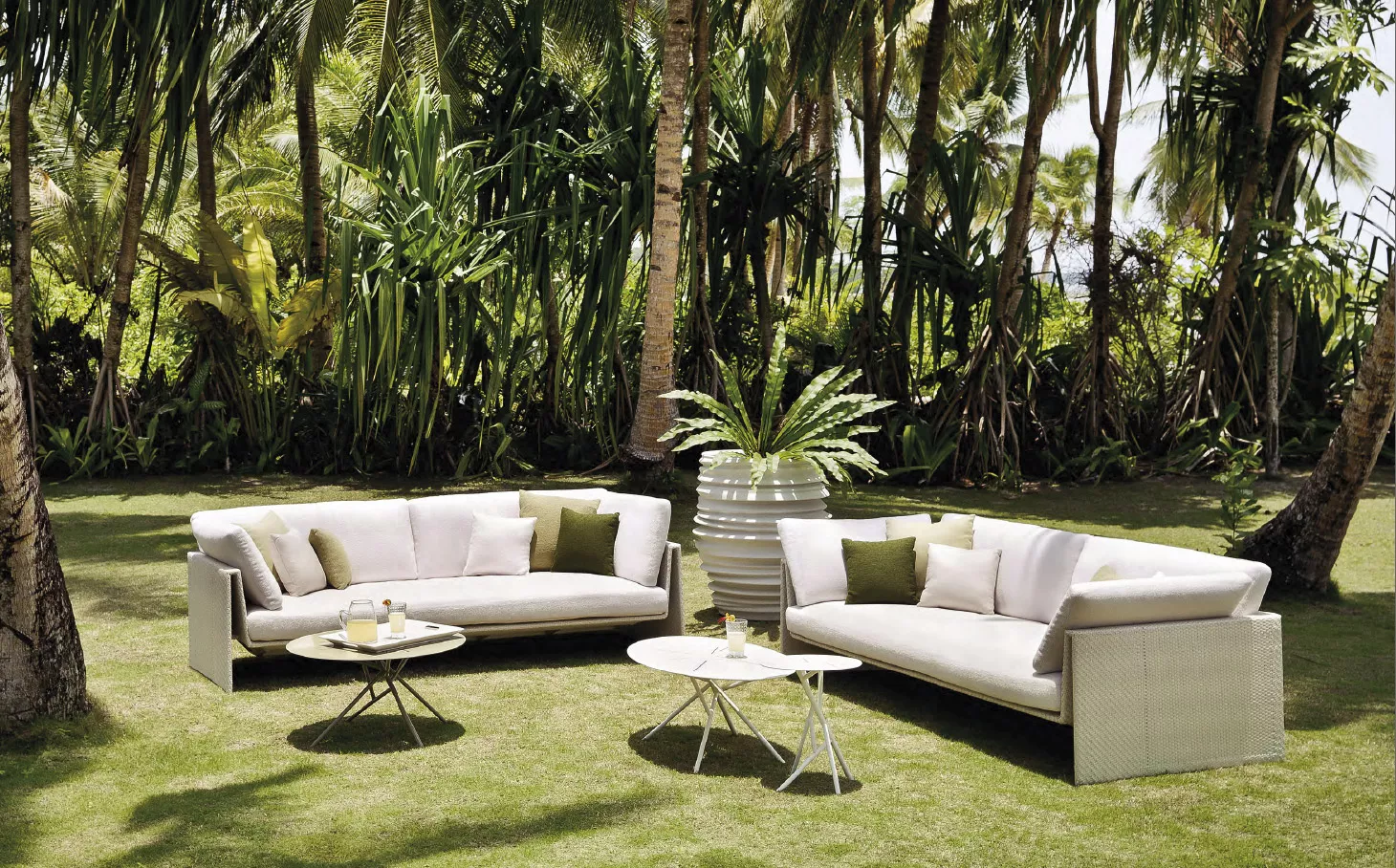 Dedon Slim Line Kollektion Dedon Panama Gartenmöbel auf einer Wiese mit Palmen im Hintergrund zwei Sofas und zwei Tische mit Getränken und Pflanzen