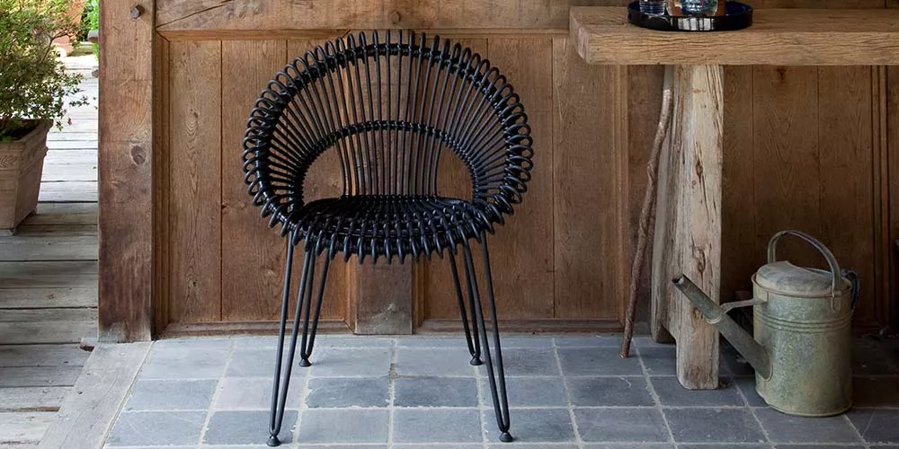 Vincent Sheppard Roxy Gartenstuhl in Schwarz vor Holzwand auf Steinboden, stimmungsvolles Bild mit rustikalem Hintergrund.