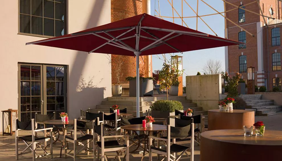 Roter Teleskopschirm von Weishäupl, rund, 500 cm, auf einer Terrasse mit Stühlen und Tischen bei Sonnenuntergang.