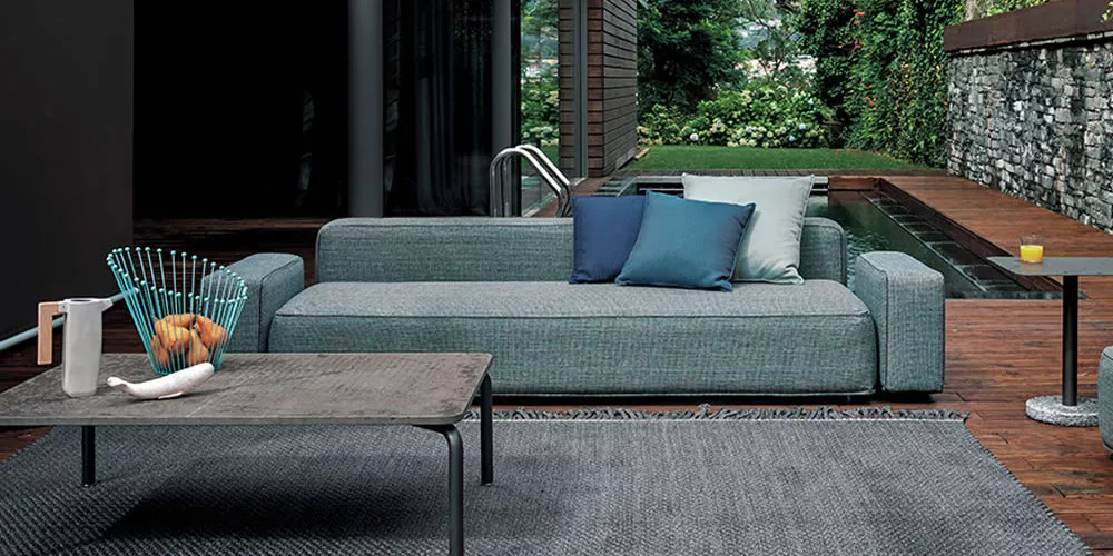 Grau-blaues Roda Dandy Sofa auf Holzterrasse mit modernem Tisch und Pool im Hintergrund, stimmungsvolle Gartenatmosphäre.