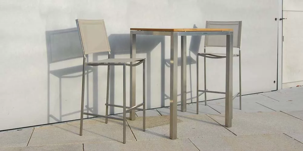 Zwei Royal Mirage New York Barhocker in Grau neben einem quadratischen Tisch auf einer Terrasse vor einer weißen Wand.