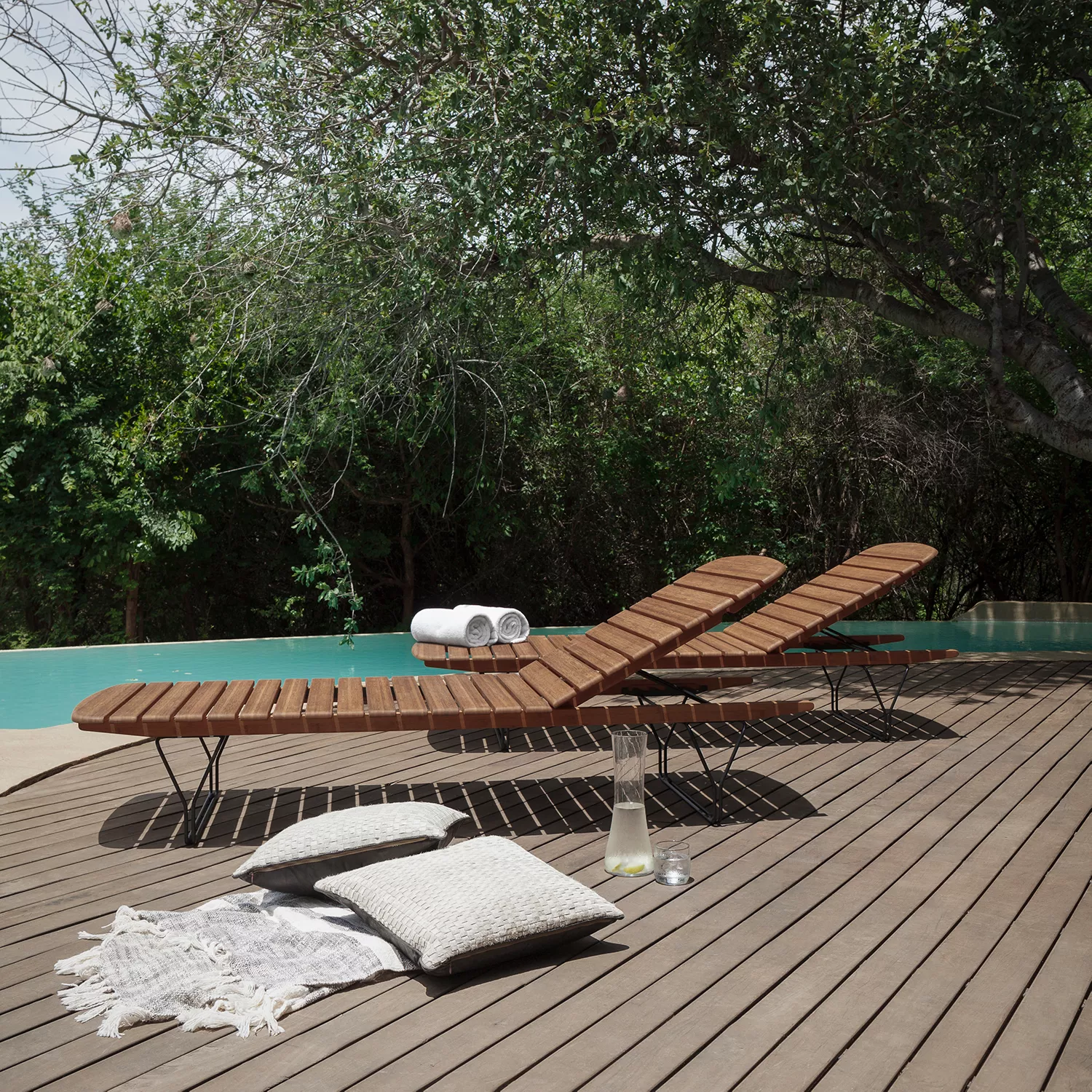 Zwei Houe Molo Sonnenliegen aus Bambus auf einer Holzterrasse neben einem Pool, umgeben von grünen Bäumen, mit Kissen und Handtüchern.