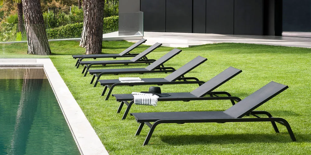 Sechs schwarze Roda Surfer Sonnenliegen auf einer grünen Rasenfläche neben einem Pool, mit Bäumen im Hintergrund.