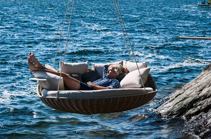 Ein Mann entspannt sich in einem Dedon Swingrest Hanging Lounger über dem Wasser an einem sonnigen Tag mood picture.