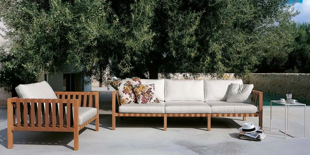 Roda Mistral Loungegruppe in Naturholz mit cremefarbenen Kissen, auf einer Terrasse vor einem großen Baum, mit Beistelltisch und Zeitschriften.