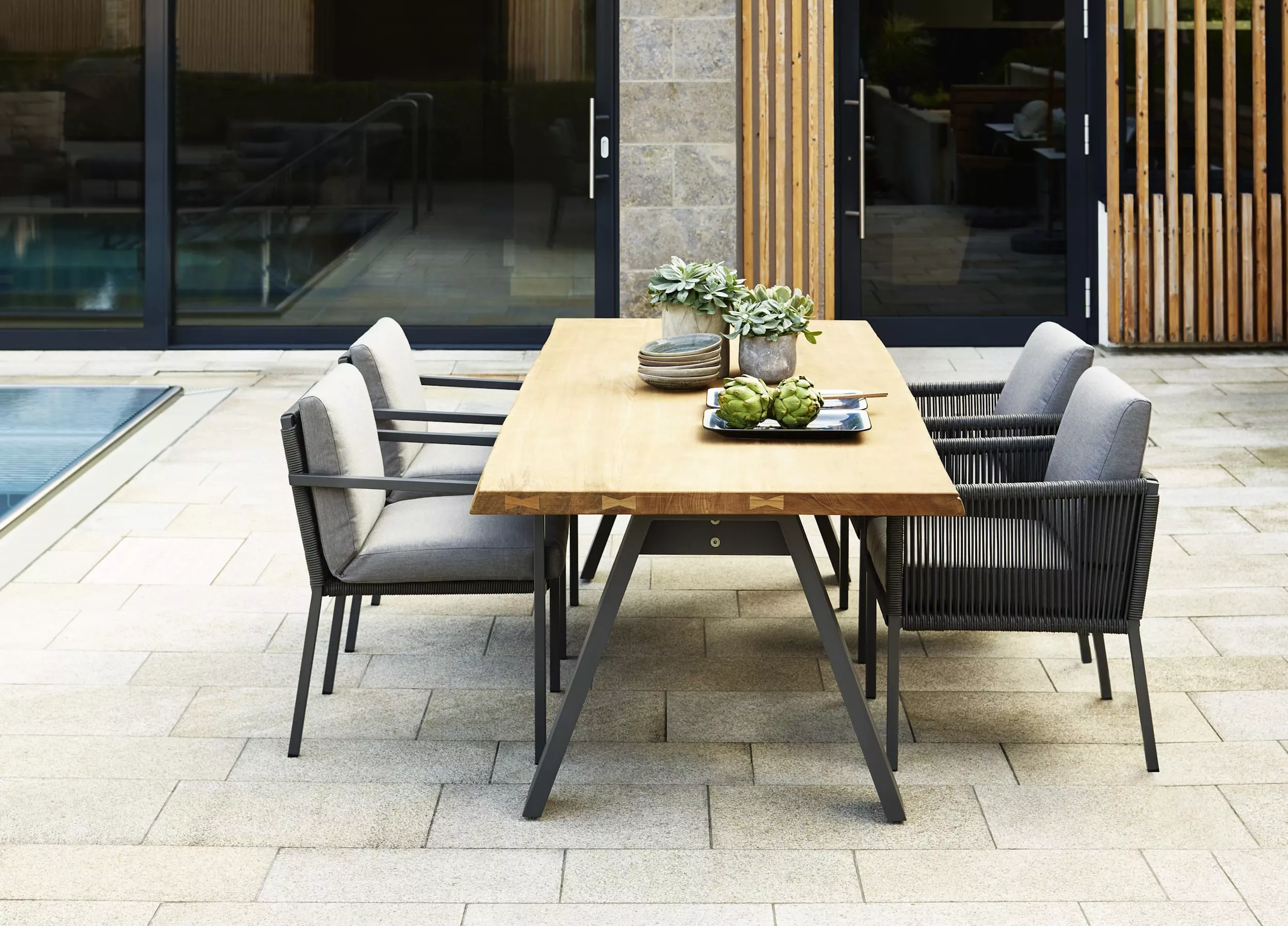 Solpuri Club Ambiente Timber Esstisch mit vier Stühlen auf einer Terrasse neben einem Pool dekoriert mit Pflanzen und Geschirr Moodbild