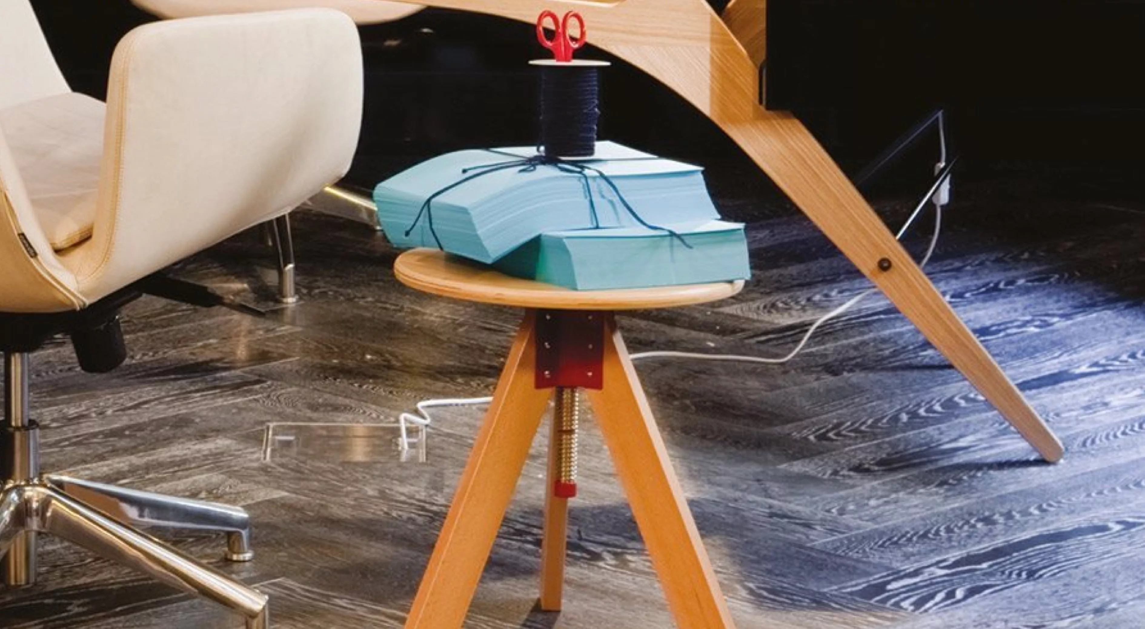 Ein Giotto-Hocker in hellem Holz auf dunklem Holzboden, mit blauen Büchern und roter Schere, in einem modernen Büro.