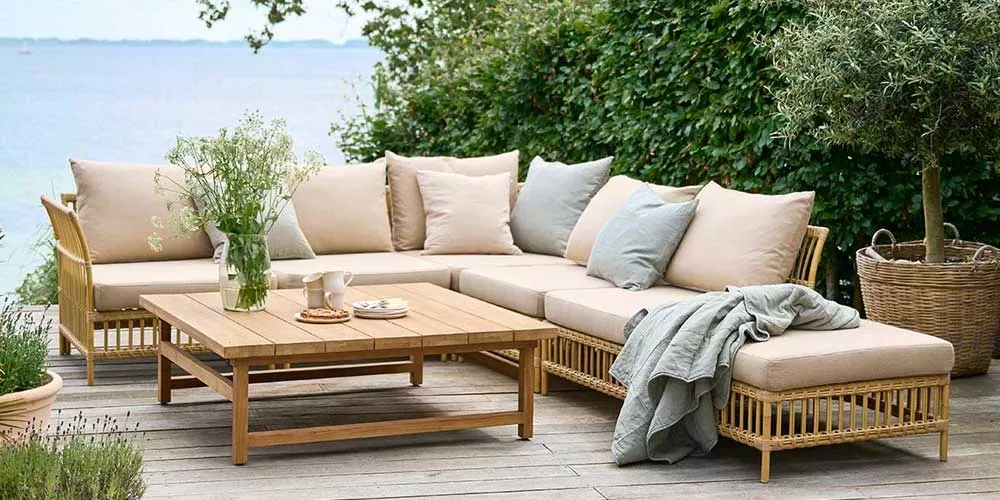 Sika-Design Exterior Loungegruppe in Beige auf Holzterrasse mit Meerblick, umgeben von grüner Hecke und Pflanzen, stimmungsvolles Ambiente.