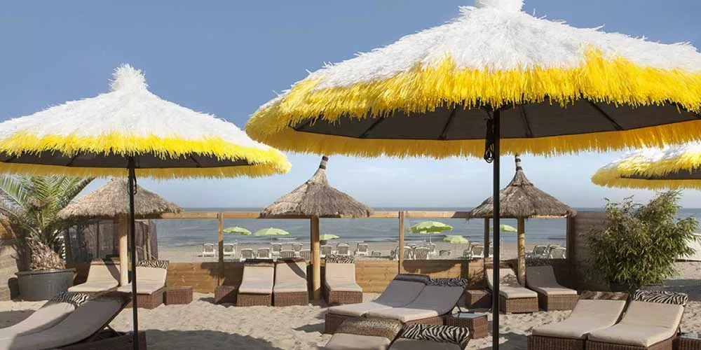 Sywawa Tahiti Sonnenschirme in Gelb und Weiß am Strand, umgeben von Liegestühlen und Palmen, mit Blick auf das Meer.