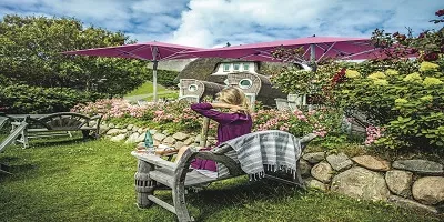 Eine Frau sitzt in einem Garten unter pinken Sonnenschirmen, umgeben von Blumen und einem Reetdachhaus im Hintergrund.