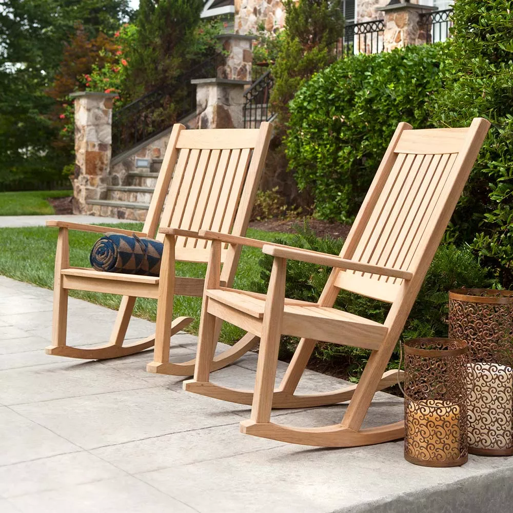Zwei Alexander Rose Roble Schaukelstühle aus hellem Holz auf einer Terrasse mit grüner Gartenkulisse und dekorativen Laternen.