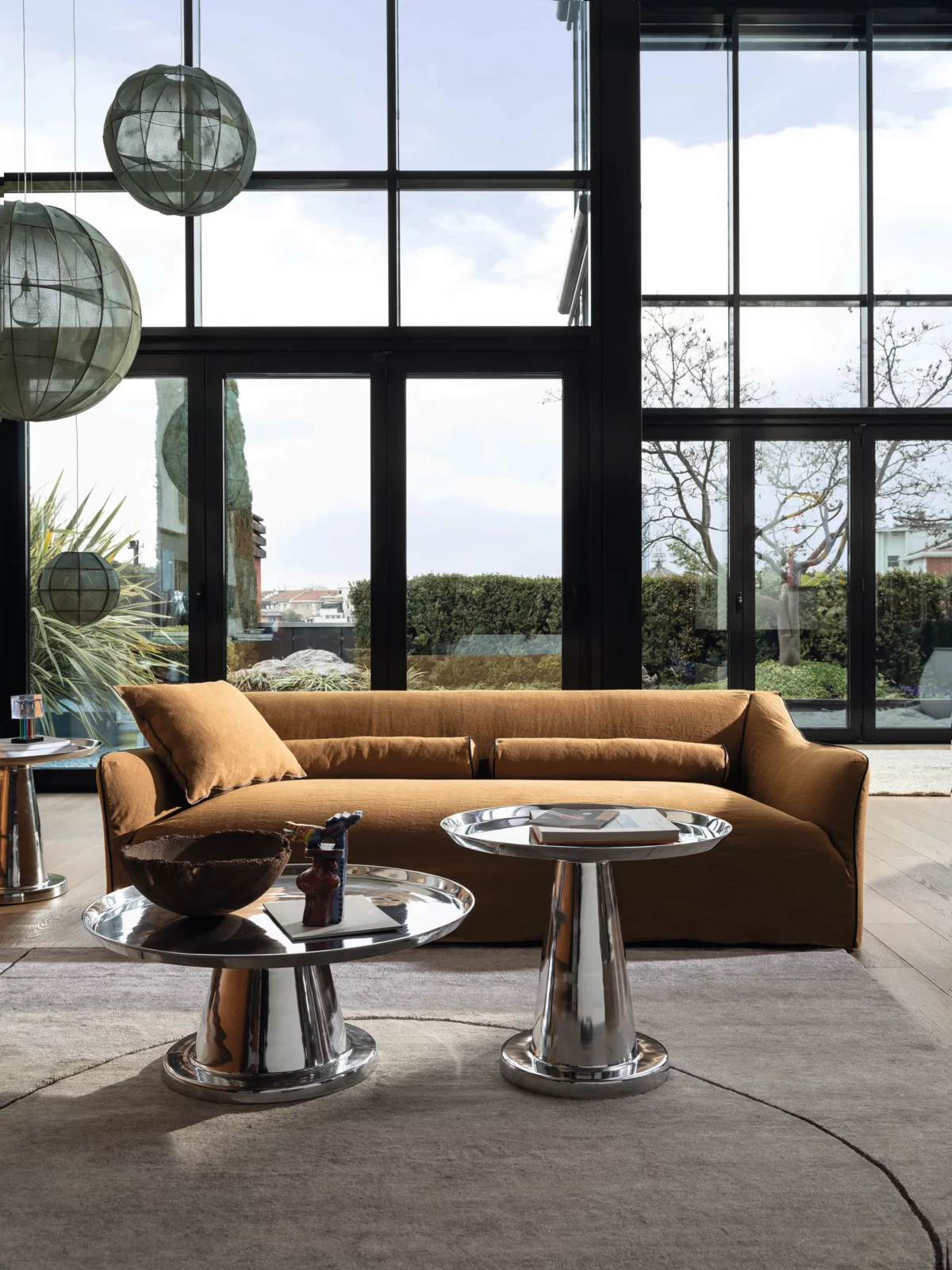 Gervasoni Sofa in einem modernen Wohnzimmer mit großen Fenstern und Blick auf den Garten mit zwei runden Beistelltischen und Hängelampen