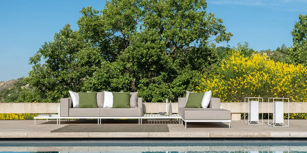 Talenti Kollektion Cleo Alu in Beige auf einer Terrasse mit grünen Kissen, umgeben von Bäumen und gelben Blumen, vor einem blauen Himmel.