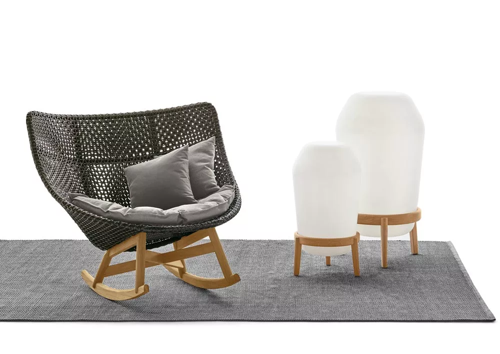 Dedon Loon Gartenleuchte und Bodenleuchte auf grauem Teppich neben einem geflochtenen Schaukelstuhl mit Kissen in einer gemütlichen Szene.