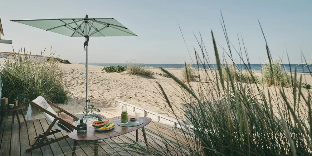 Glatz Forntino Sonnenschirm in Mintgrün auf einer Holzterrasse am Strand mit Liegestuhl und Tisch, sommerliche Strandatmosphäre.