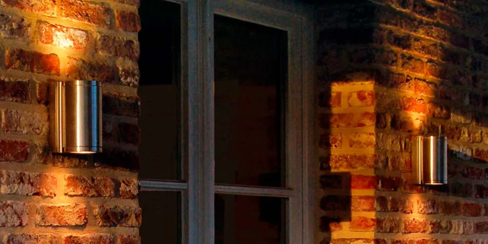 Royal Botania Omega Gartenleuchten in Edelstahl an einer Backsteinwand, stimmungsvolle Beleuchtung bei Dämmerung.