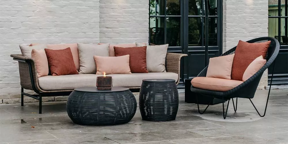 Vincent Sheppard Ivo Gartenmöbel-Set in Beige und Rot auf einer Terrasse vor einer weißen Backsteinwand, bestehend aus Sofa, Sessel und Tischen.