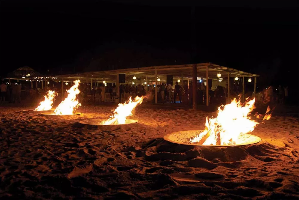 Drei AK47 Hole Feuerstellen aus oxidiertem Stahl brennen am Strand vor einem beleuchteten Pavillon bei Nacht.