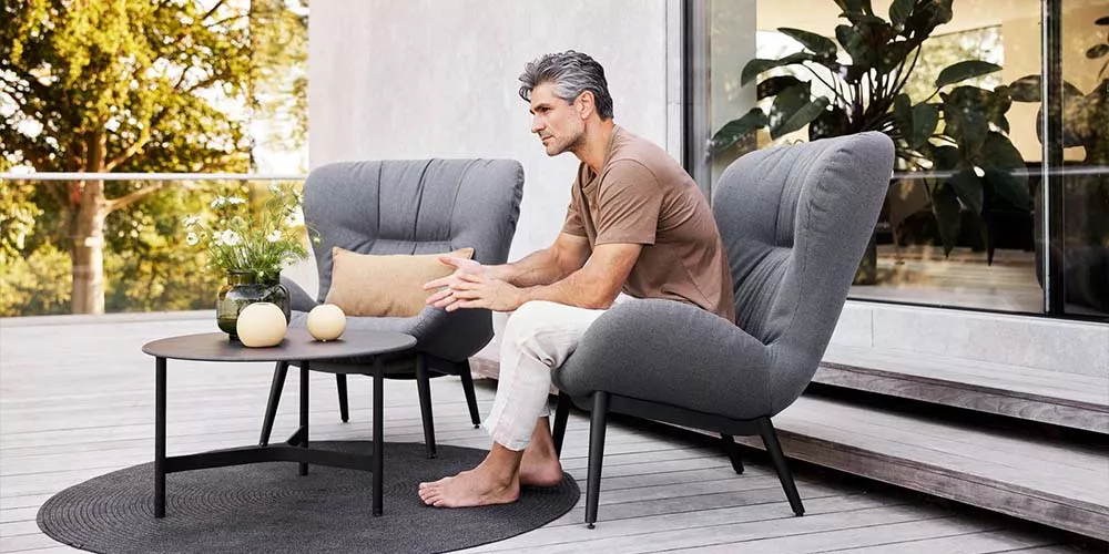 Ein Mann sitzt auf einem grauen Caneline Serene Sessel auf einer Terrasse, umgeben von Pflanzen und einem runden Tisch mit Dekoration.