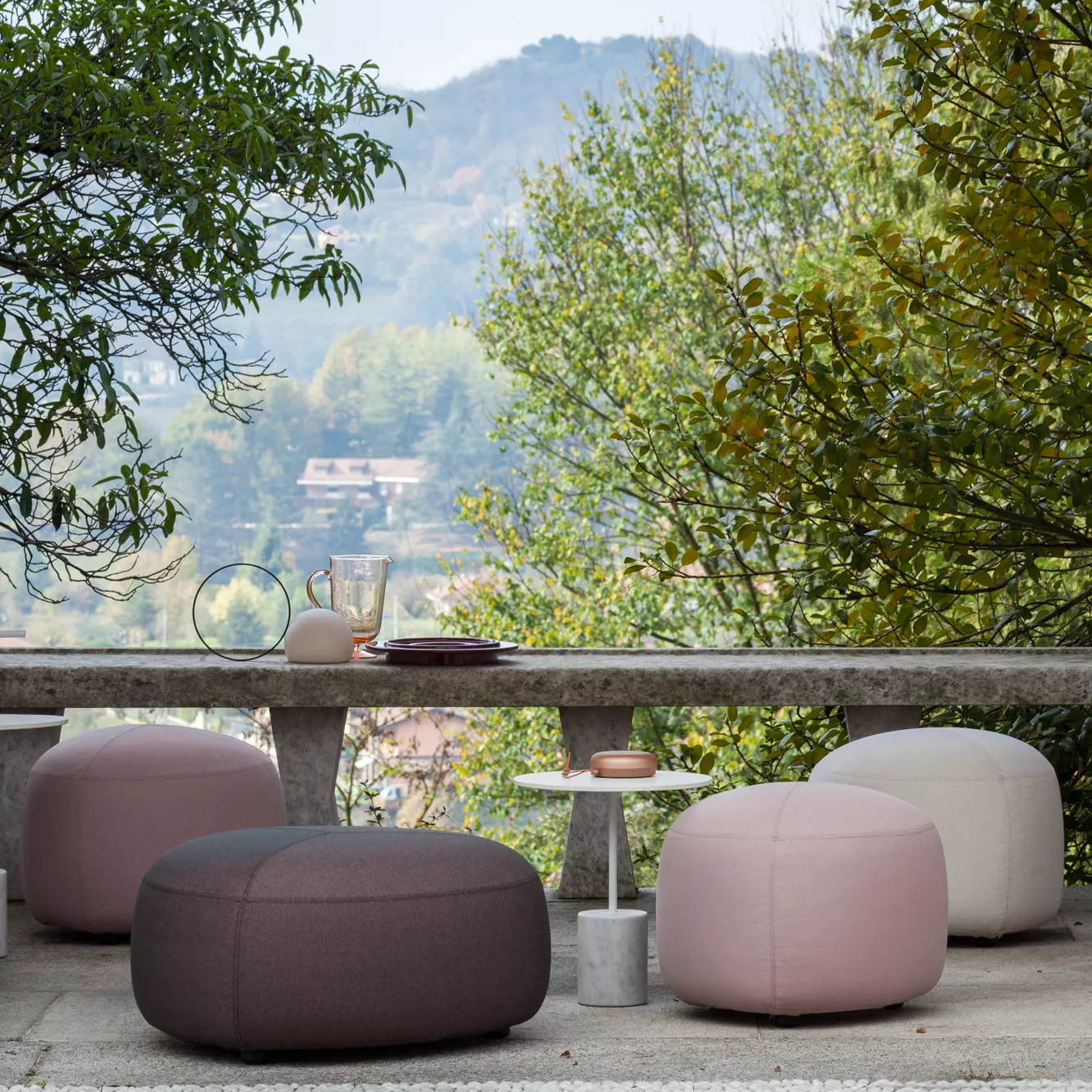 Lapalma Kipu Hocker in Rosa und Grau auf einer Terrasse mit Naturhintergrund, stimmungsvolles Outdoor-Setting.