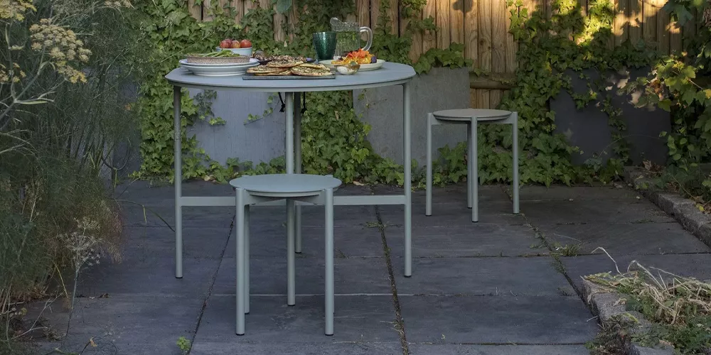 Skagerak Picnic Tisch und Hocker in Hellgrau auf einer Terrasse mit Pflanzen im Hintergrund, gedeckt mit Geschirr und Speisen.